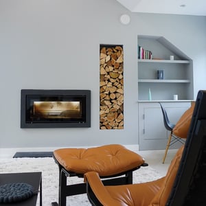 brown foot stool in foreground with log burner behind and logs stacked in wall reveal